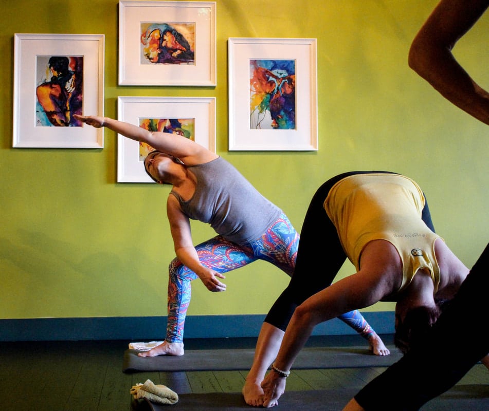 "In a mysore style Ashtanga Yoga class led by Krista Shirley, two students are depicted fully engaged in their practice. One student gracefully executes Prasarita Padottanasana, while the other is in a modified version of Side Angle Pose. Krista, a Level Two authorized Ashtanga Yoga instructor, emphasizes traditional principles in her teaching at The Yoga Shala in Maitland, Florida. The studio, located at 140 Circle Drive #4, specializes in the renowned Mysore method, providing transformative experiences for students both in-person and virtually. Krista shares her expertise through her YouTube channel, enriching the yoga journeys of practitioners worldwide."