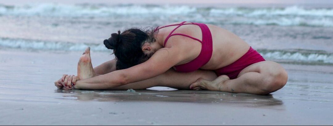 Krista Shirley, a level 2 authorized Ashtanga Yoga teacher and owner of the Yoga Shala in Maitland, Florida, is seated on the beach in the water, performing the Ashtanga Yoga posture Janu Sirsasana A. She exudes tranquility and concentration, dressed in professional yoga attire. The serene backdrop of the beach sand enhances the peaceful ambiance.
